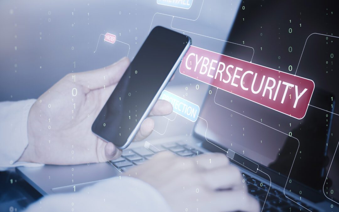 A hand using a mobile and laptop, with the words ‘cybersecurity’ displaying on the laptop's screen